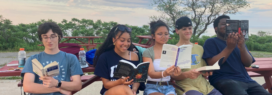 Five students sit side by side on a park bench reading books.