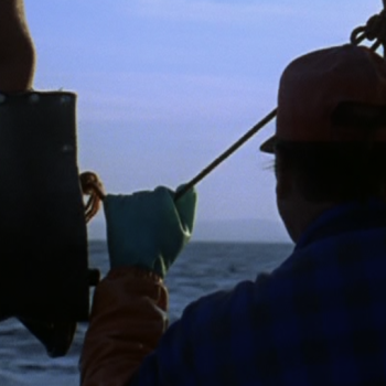 Closeup of man facing away from the camera on a fishing boat. He is silhouetted against a blue-purple sky and dark blue water.