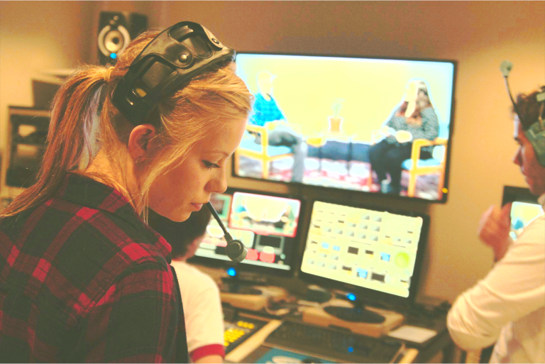 Students in Intro to Video Production stand in control room. A young woman wears a headset and is in front of a computer screen. In the background through a window is a person sitting in a chair.