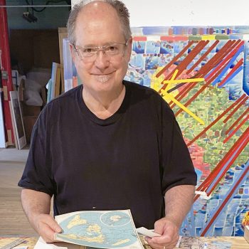 Photograph of artist Dan Mills in his art studio. Dan stands behind a work table with maps in his hands. One of his original paintings is directly behind him and he is pictured in an open building. Dan faces the camera with a smile.