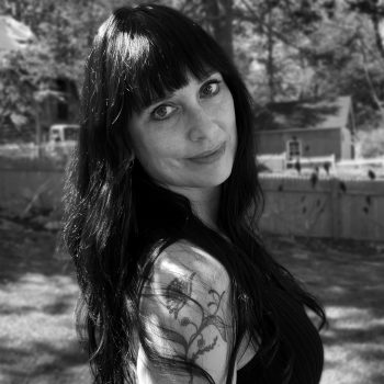 Black and white photograph of artist and author, Summer J. Hart. Summer stands in front of a natural background, facing the camera and looking over her shoulder.