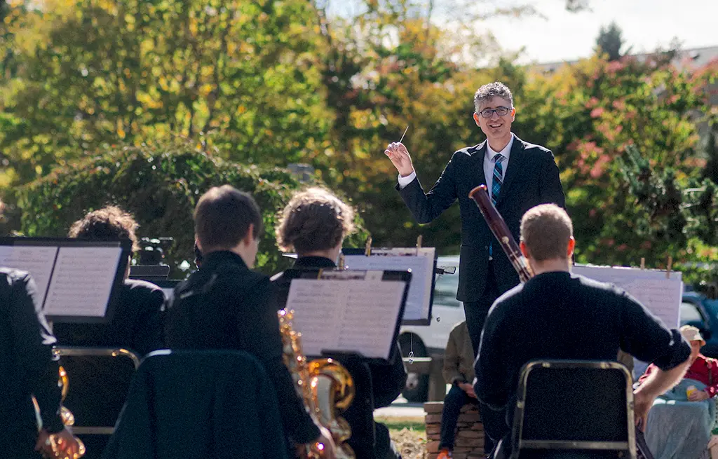 A university conductor smiles at the camera during an outdoor band concert.