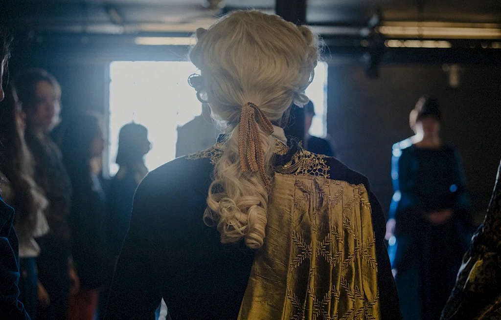 A theatre student wearing a powdered wig and period clothing faces a window.