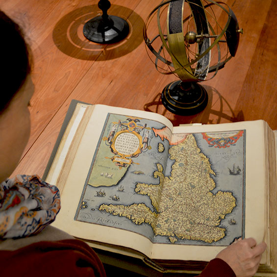 A student flips through a old book of maps which is open on a wooden table next to antique globes.