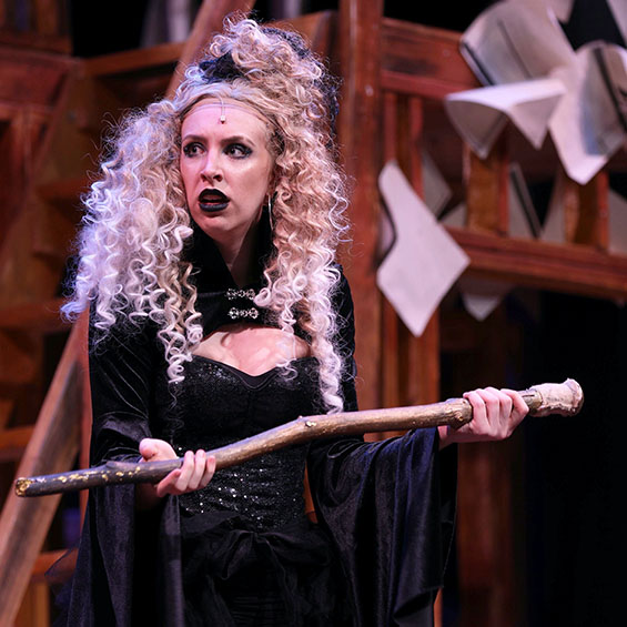Kallie Brown performs on stage as the witch in our spring production of 'Into the Woods.' Dressed in all black with a blond wig, she holds up a wooden walking stick.
