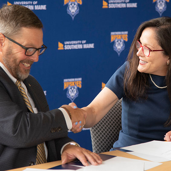 SMCC President Joe Cassidy and our President Jacqueline Edmondson shake hands at the signing of the Southern Maine Pathways Agreement. A dark blue backdrop behind them features the University of Southern Maine logo and our Southern Maine Huskies graphic.