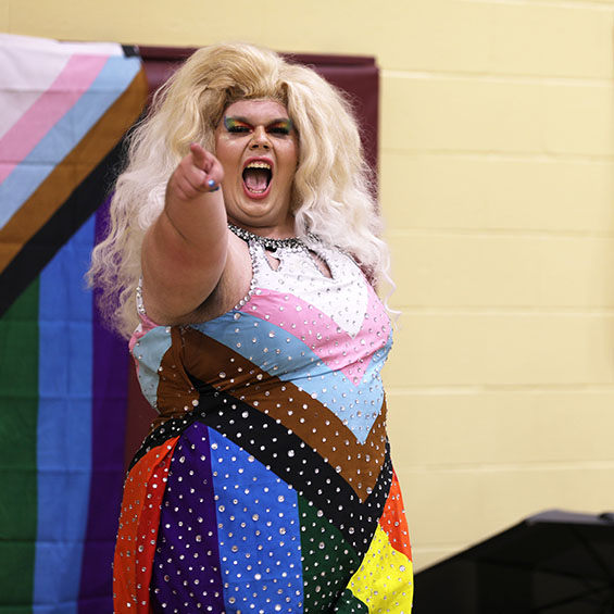 Queen Letta, alternately known as theatre major Bryan Spaulding, points at the camera during a drag performance. An intersex-inclusive pride flag is on the gymnasium wall behind her.