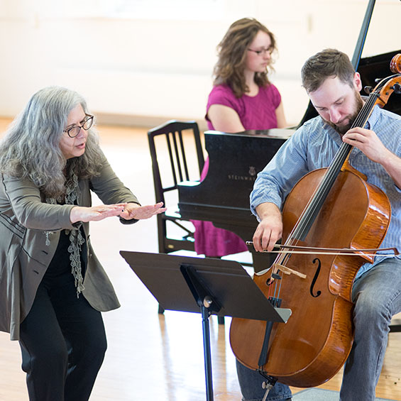 A faculty member conducts a student playing the cello in the foreground and a student playing the piano in the background during a rehearsal in Corthell Hall.
