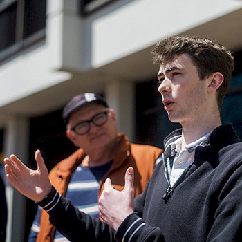 Jacob Curtis speaking animatedly to a group of people that includes faculty member Richard Bilodeau, seen in the background wearing an orange coat.