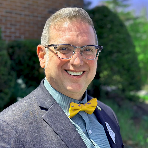 An outdoor portrait of Joshua Chard. He's wearing glasses, a yellow bowtie, and a grey blazer. Photo courtesy of the Maine Department of Education