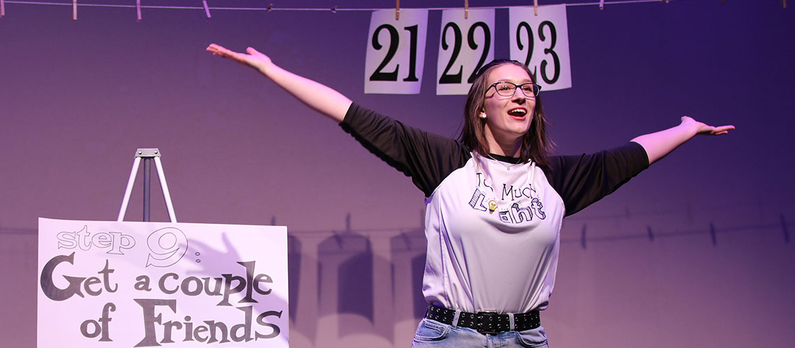 A student actor wearing a baseball-style t-shirt that reads 'Too Much Light' performs with their arms stretched out. A sign in the background reads, 'Step 9: Get a couple of friends.' The numbers 21, 22, 23 are printed large on individual sheets of 8.5 x 11 inch paper and pinned to a clothesline that across the entire stage.