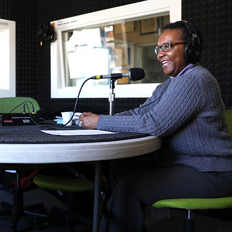 Dr. Idella Glenn in a recording studio at WMPG. She's wearing over-the-ear headphones and speaking into a mic mounted on the table in front of her.