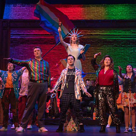 Student actors on stage during a performance of "The Prom." One student is dressed as the Statue of Liberty, and another is waving a pride flag.