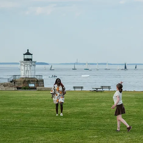 Two students play frisbee on the grass by Bug Light lighthouse.