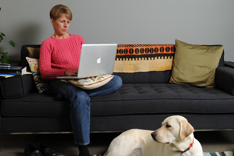Person sits on a couch with a computer in their lap and a dog curled up on the floor.