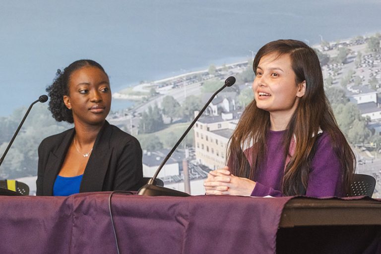 Two professionals seated behind a table with microphones speaking at a panel.