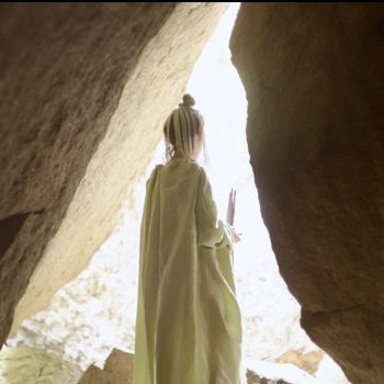 A woman dressed in a green robe stands at a cave opening. Light pours in through the crevice. Her hair is in a bun on the top of her head and she wears a sheer veil.
