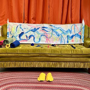 A yellow couch with a scroll of abstract art sppread across its back. At left, several bolts of rolled fabric. In the center, in front of the couch, is a pair of bright yellow sneakers sitting on a striped oval rug.