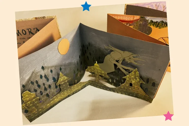 A handmade book is open to reveal a creature creeping behind a village — "Kikimora Mythology" by Kathleen Bender