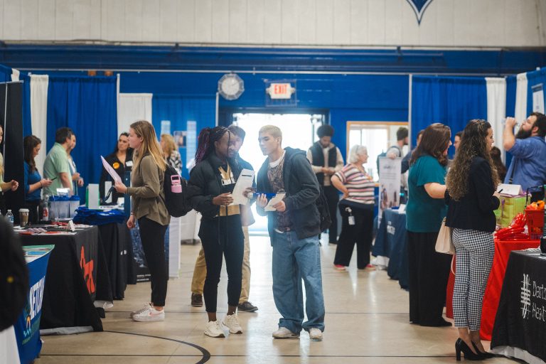 Students are milling around tables lined up along the sides, speaking with employers in a gym.
