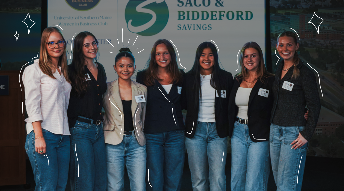 Seven students stand together in front of a University of Southern Maine Women in Business Club banner and a Saco and Biddeford Savings sign at a networking event.