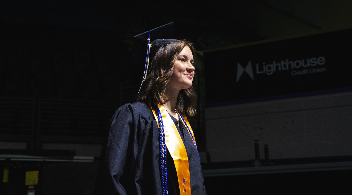 A graduate in a cap, gown, and gold USM stole stands in the staging area at Cross Insurance Arena, looking ahead before the processional begins.