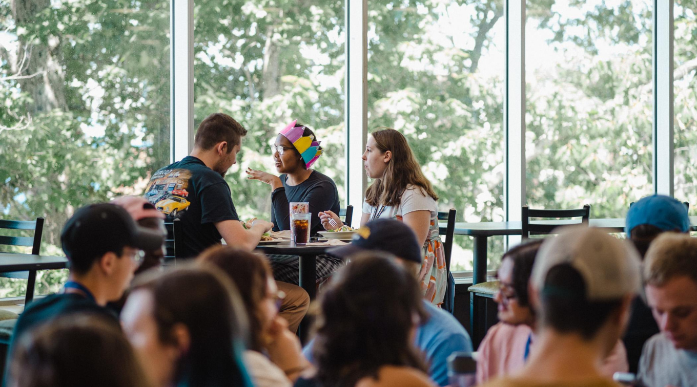 Students chat over lunch at a table by large windows in a busy campus dining hall.