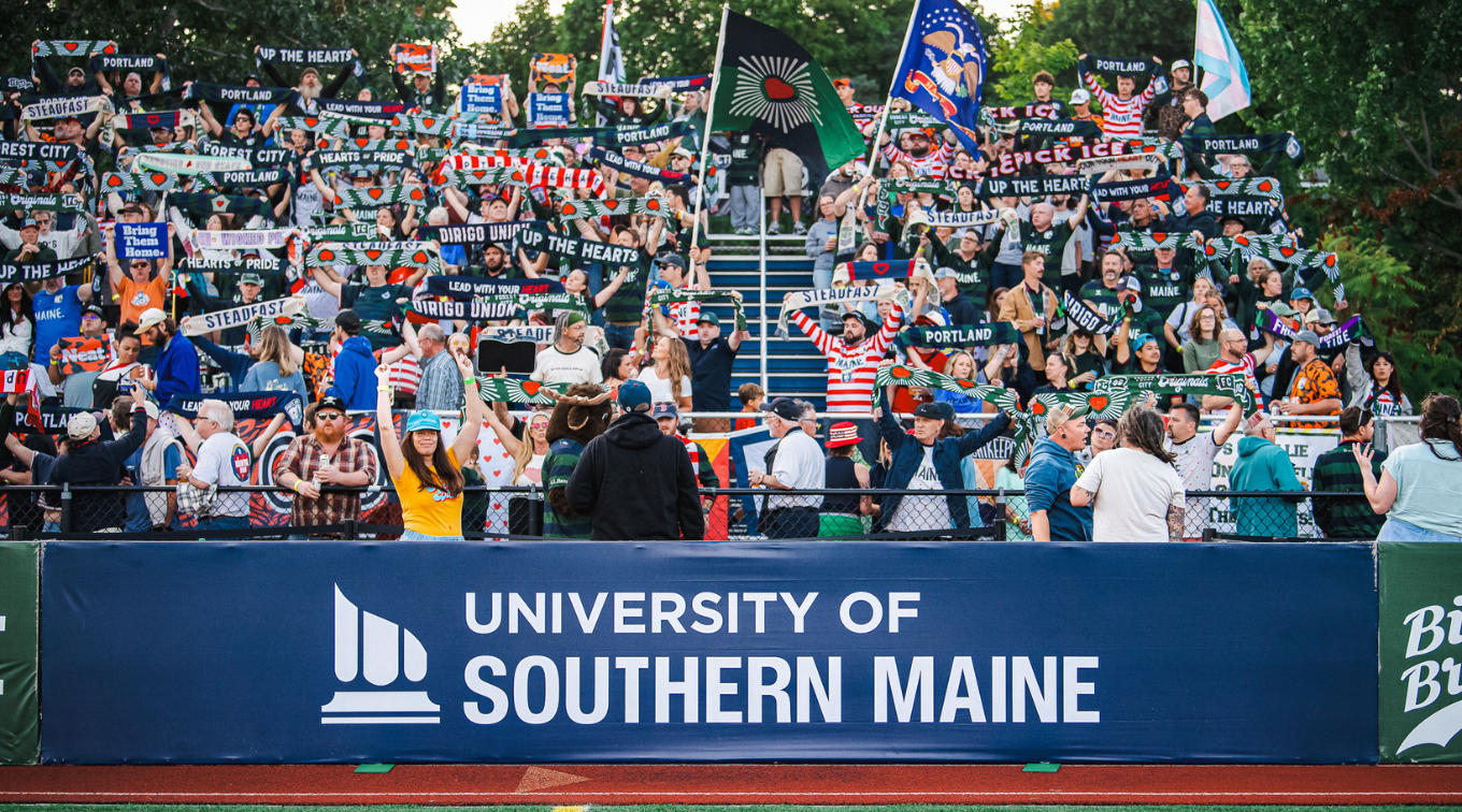 Hearts of Pine fans wave scarves and flags behind a University of Southern Maine banner at Fitzpatrick Stadium.